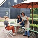 Twee vrouwen zitten aan een buitentafel bij een café terwijl een lachende serveerster hen bedient.
