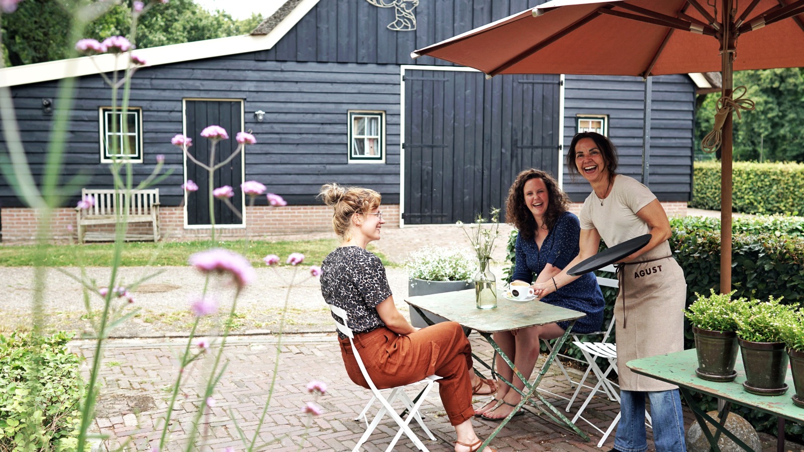 Twee vrouwen zitten aan een buitentafel bij een café terwijl een lachende serveerster hen bedient.