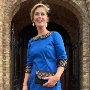 Vrouw in een blauwe jurk met bloemenaccenten staat lachend voor een stenen boog en een zwart hek.