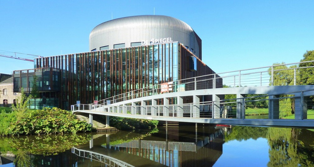 Modern gebouw met grote ramen genaamd 'De Spiegel', brug en reflectie in het water bij zonnig weer.
