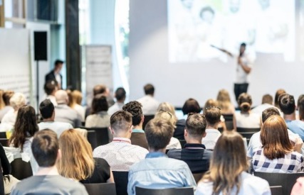 A group of people attending a presentation or seminar, listening to a speaker in front of a projection screen.