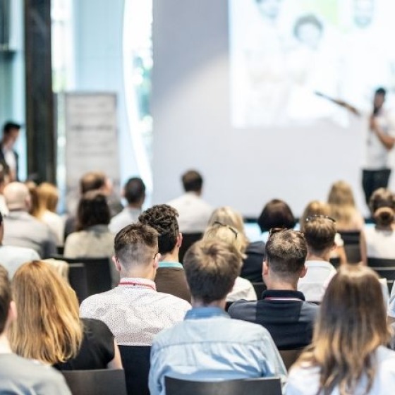 Eine Gruppe von Menschen sitzt in einem Raum und hört einem Redner zu, der eine Präsentation hält.
