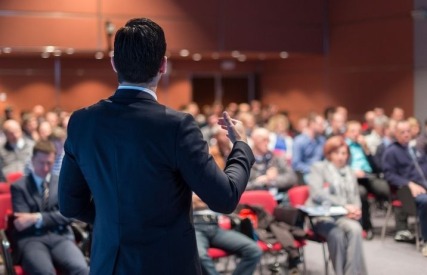 A man in a suit delivers a presentation to an attentive audience seated in a conference room.