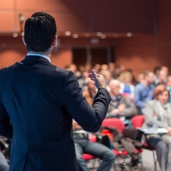 Ein Mann im Anzug hält eine Präsentation vor einem aufmerksamen Publikum in einem Konferenzraum.