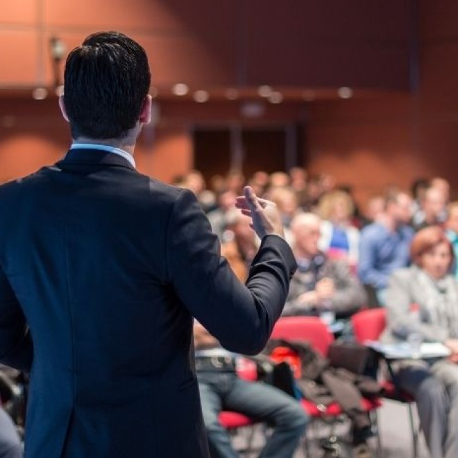 Een man in pak geeft een presentatie voor een geïnteresseerd publiek in een conferentiezaal.