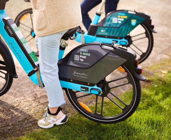 Zwei Personen stehen mit blauen E-Bikes zum Teilen auf einem Weg neben Gras, bereit für eine gemeinsame Fahrt.