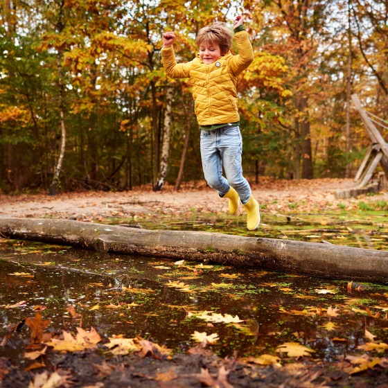 Een kind in een gele jas springt over een waterplas in een herfstig bos, omringd door gekleurde bladeren.