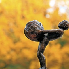 Bronzeskulptur einer Person, die eine große Kugel trägt, vor einem unscharfen Hintergrund aus Herbstlaub.