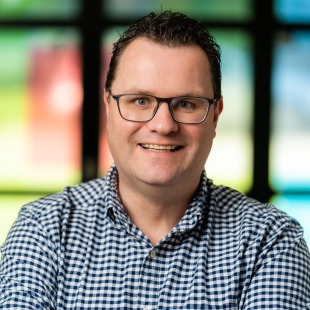 Portret van een glimlachende man met bril en ruitjesoverhemd voor een kleurrijke, wazige achtergrond.