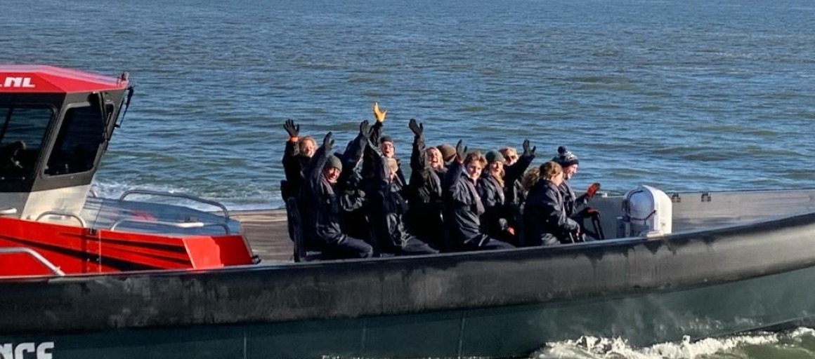 Een groep mensen in zwarte kleding vaart op een speedboot over zee, zwaaiend naar de camera.