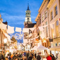Drukke kerstmarkt in een sfeervolle Nederlandse straat met lichtjes en historische gebouwen bij schemering.