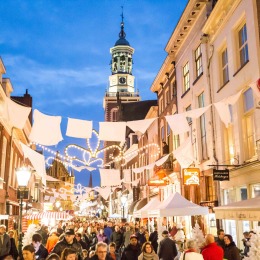 Belebter Weihnachtsmarkt in einer stimmungsvollen niederländischen Straße mit Lichtern und Kirchturm am Abend.