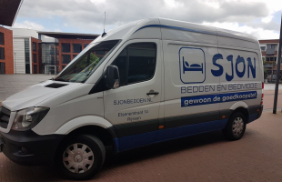 Bestelwagen met opdruk van SJON bedden en bedmode, geparkeerd op straat in een stedelijk gebied.