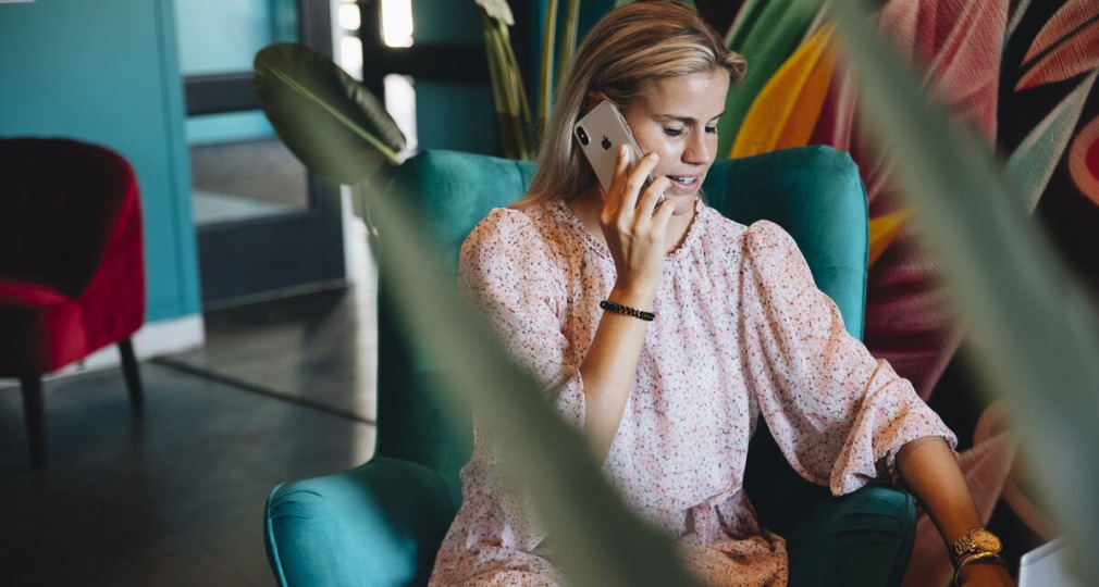 Vrouw met een lichtroze jurk belt en werkt op een laptop terwijl ze in een moderne lounge zit.