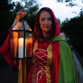 Vrouw in rood-groene cape met een lantaarn, staand buiten tijdens schemering op een houten brug.