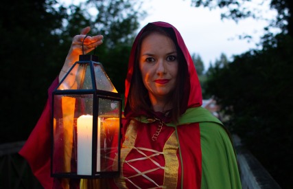 Vrouw in rood-groene cape met een lantaarn, staand buiten tijdens schemering op een houten brug.
