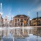 Fountains shoot up on a square with historic buildings in the background and a person walking by.