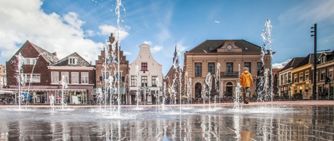 Fonteinen spuiten omhoog op een plein met historische gebouwen op de achtergrond en een wandelaar.