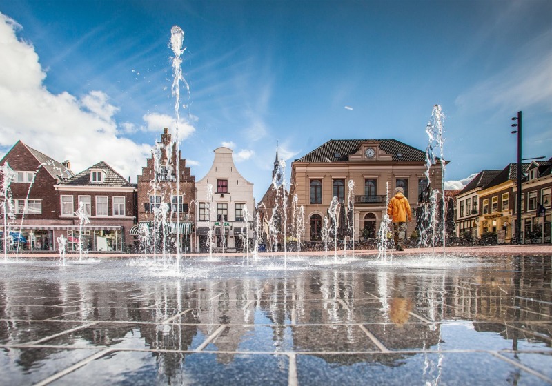 Fonteinen spuiten omhoog op een plein met historische gebouwen op de achtergrond en een wandelaar.