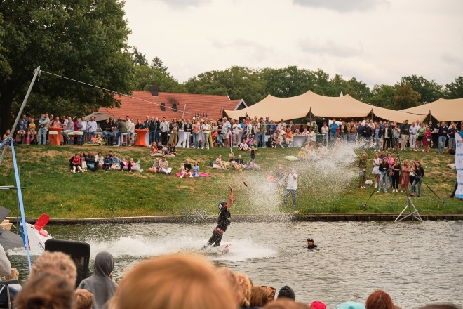 Wakeboarder voert stunt uit op water tijdens evenement, omringd door publiek aan de waterkant.
