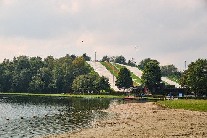 Strand, meer en een kunstmatige skihelling met groene en witte banen in een bosrijk park in de zomer.
