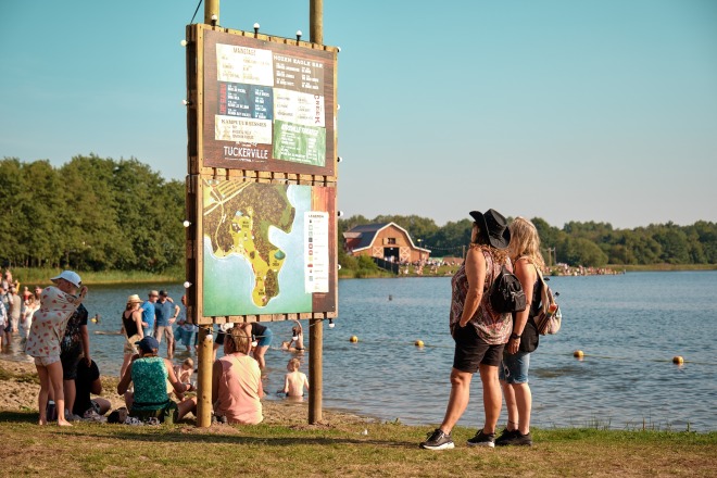 Mensen bekijken een festivalplattegrond bij het water, met een boerderij en bezoekers op de achtergrond.