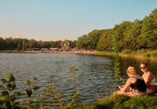 Twee vrouwen zitten aan de oever van een meer met op de achtergrond een kermis en veel mensen.