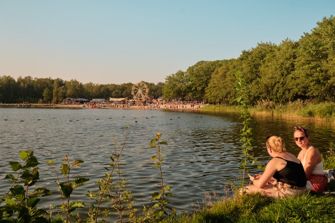 Twee vrouwen zitten aan de oever van een meer met op de achtergrond een kermis en veel mensen.