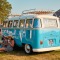 Mensen genieten van een zomers festival, zittend op het gras naast een blauwe Volkswagen bus.