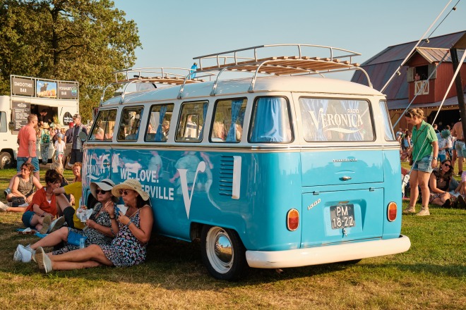 Mensen genieten van een zomers festival, zittend op het gras naast een blauwe Volkswagen bus.