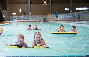 Kinderen leren zwemmen met drijfborden in een overdekt zwembad onder begeleiding van een instructeur.