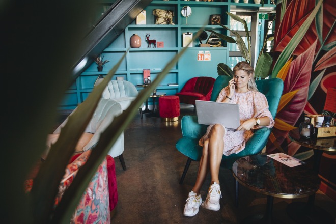 Vrouw werkt op een laptop en telefoneert in een kleurrijke, moderne lounge met planten en kunst.