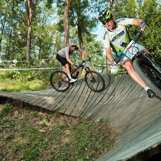 Twee mountainbikers rijden snel over een houten bocht in een bosrijk parcours op een zonnige dag.