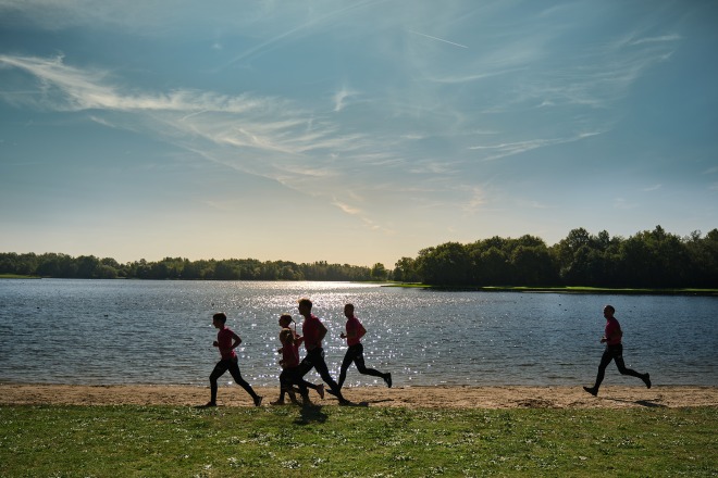 Groep mensen rent langs een meer onder een heldere lucht, omgeven door bomen en natuur, op een zonnige dag.