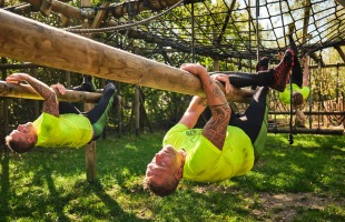 Deelnemers in gele shirts nemen deel aan een hindernisbaan en hangen aan houten balken buiten.
