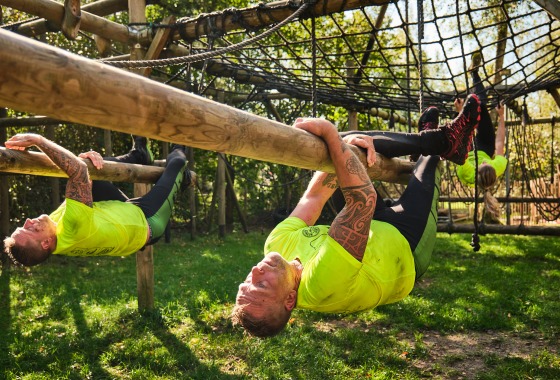 Deelnemers in gele shirts nemen deel aan een hindernisbaan en hangen aan houten balken buiten.