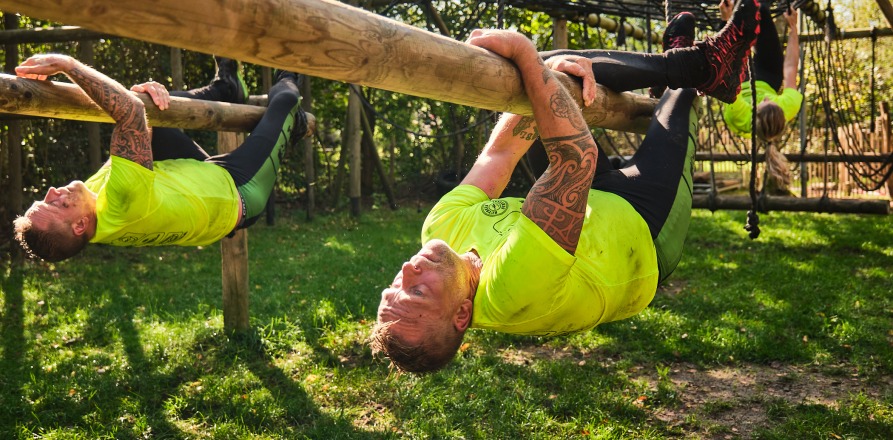 Deelnemers in gele shirts nemen deel aan een hindernisbaan en hangen aan houten balken buiten.