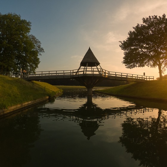 Houten brug met unieke torenoverkapping over een rustige gracht bij zonsondergang, omgeven door bomen.