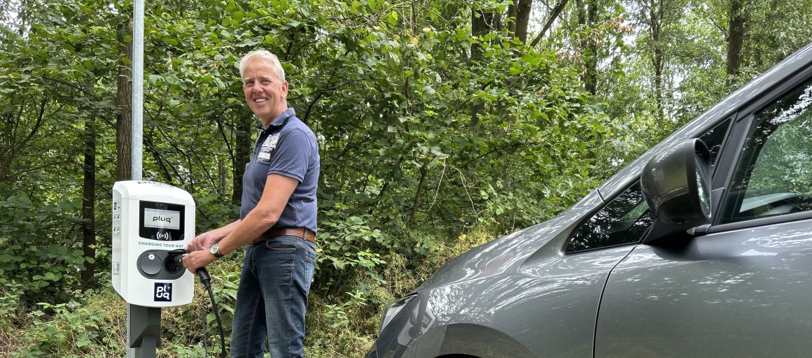 Man laadt een elektrische auto op bij een laadpaal in een bosrijke omgeving, glimlacht naar de camera.