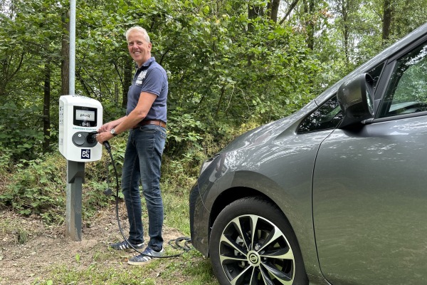 Man laadt een elektrische auto op bij een laadpaal in een bosrijke omgeving, glimlacht naar de camera.