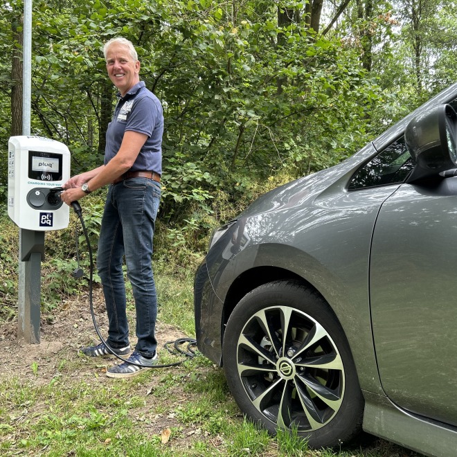 Man laadt een elektrische auto op bij een laadpaal in een bosrijke omgeving, glimlacht naar de camera.
