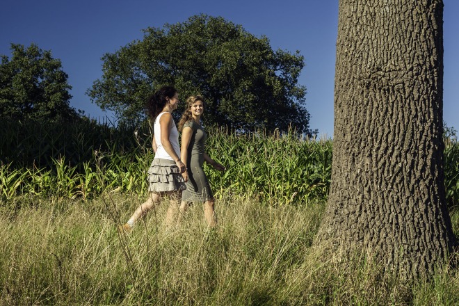Twee vrouwen wandelen in zomerse kleding langs hoog gras en bomen op een zonnige dag in de natuur.