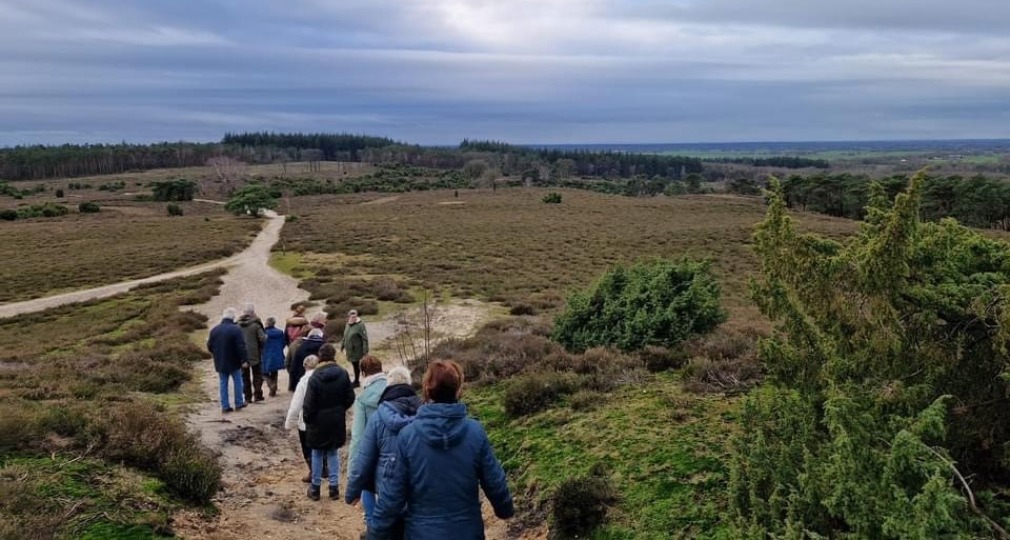 Groep mensen wandelt over een zandpad in een uitgestrekt natuurgebied met heuvels en bos op de achtergrond.