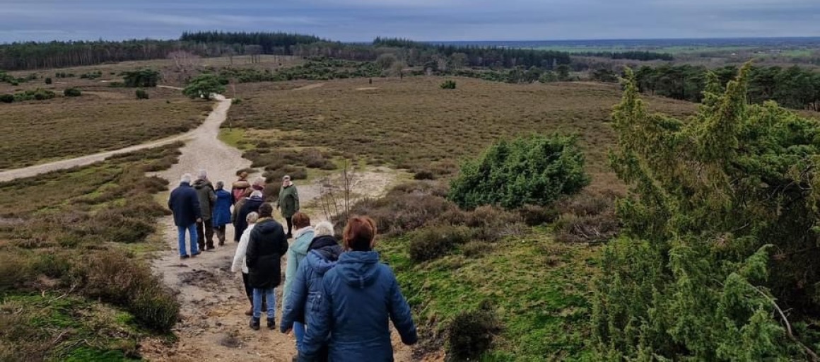 Groep mensen wandelt over een zandpad in een uitgestrekt natuurgebied met heuvels en bos op de achtergrond.