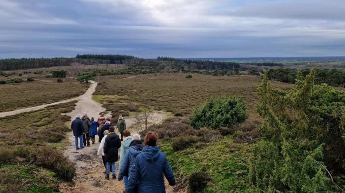 Groep mensen wandelt over een zandpad in een uitgestrekt natuurgebied met heuvels en bos op de achtergrond.
