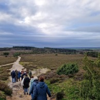 Groep mensen wandelt over een zandpad in een uitgestrekt natuurgebied met heuvels en bos op de achtergrond.