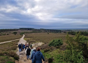 Groep mensen wandelt over een zandpad in een uitgestrekt natuurgebied met heuvels en bos op de achtergrond.