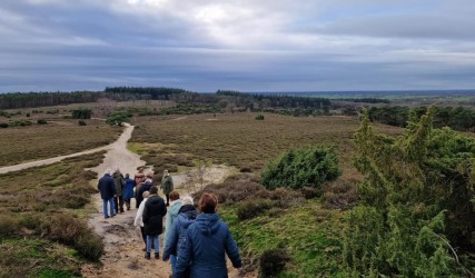 Groep mensen wandelt over een zandpad in een uitgestrekt natuurgebied met heuvels en bos op de achtergrond.