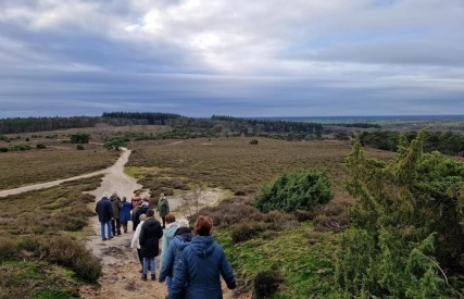 Groep mensen wandelt over een zandpad in een uitgestrekt natuurgebied met heuvels en bos op de achtergrond.