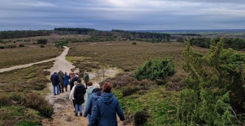Groep mensen wandelt over een zandpad in een uitgestrekt natuurgebied met heuvels en bos op de achtergrond.